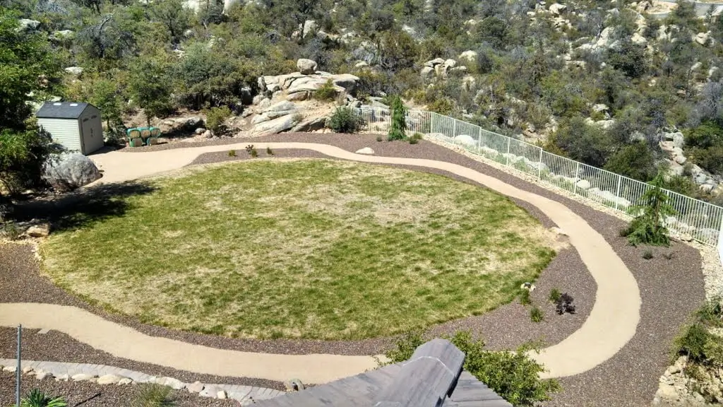 Aerial view of a circular grassy area surrounded by a paved walkway, landscaping rocks, a small shed, and a metal fence with trees and rocks in the background.