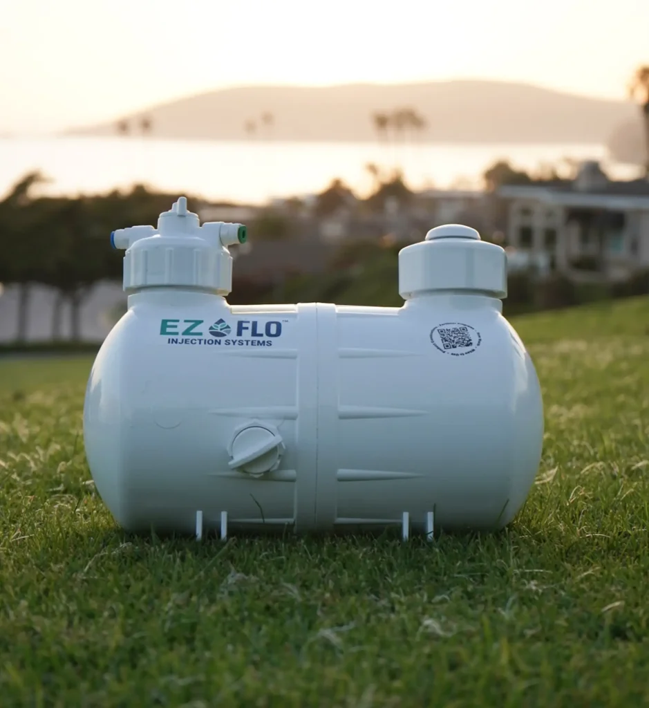 A white EZ FLO injection system tank sits on grass with a blurred background of houses, trees, and water at sunset.