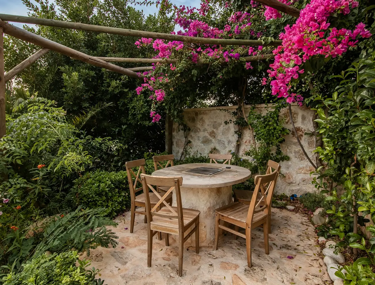 A round stone table with four wooden chairs sits on a stone patio under a wooden pergola with vibrant pink flowers and lush green plants surrounding the area.