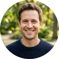 A man with short brown hair and a dark sweater smiles outdoors with greenery in the background.